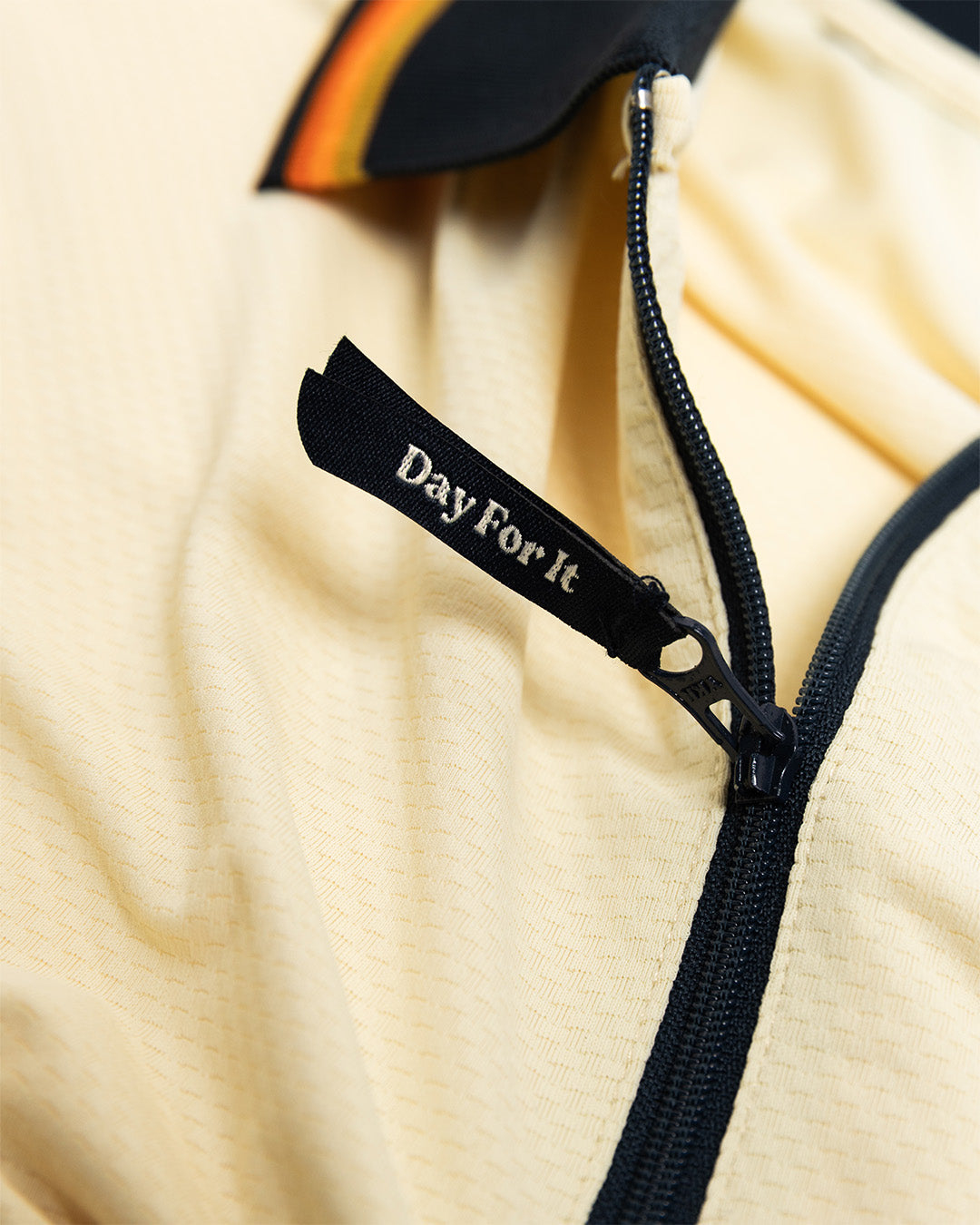 Close-up of a beige jacket with a black zipper and 'Day For It' label.