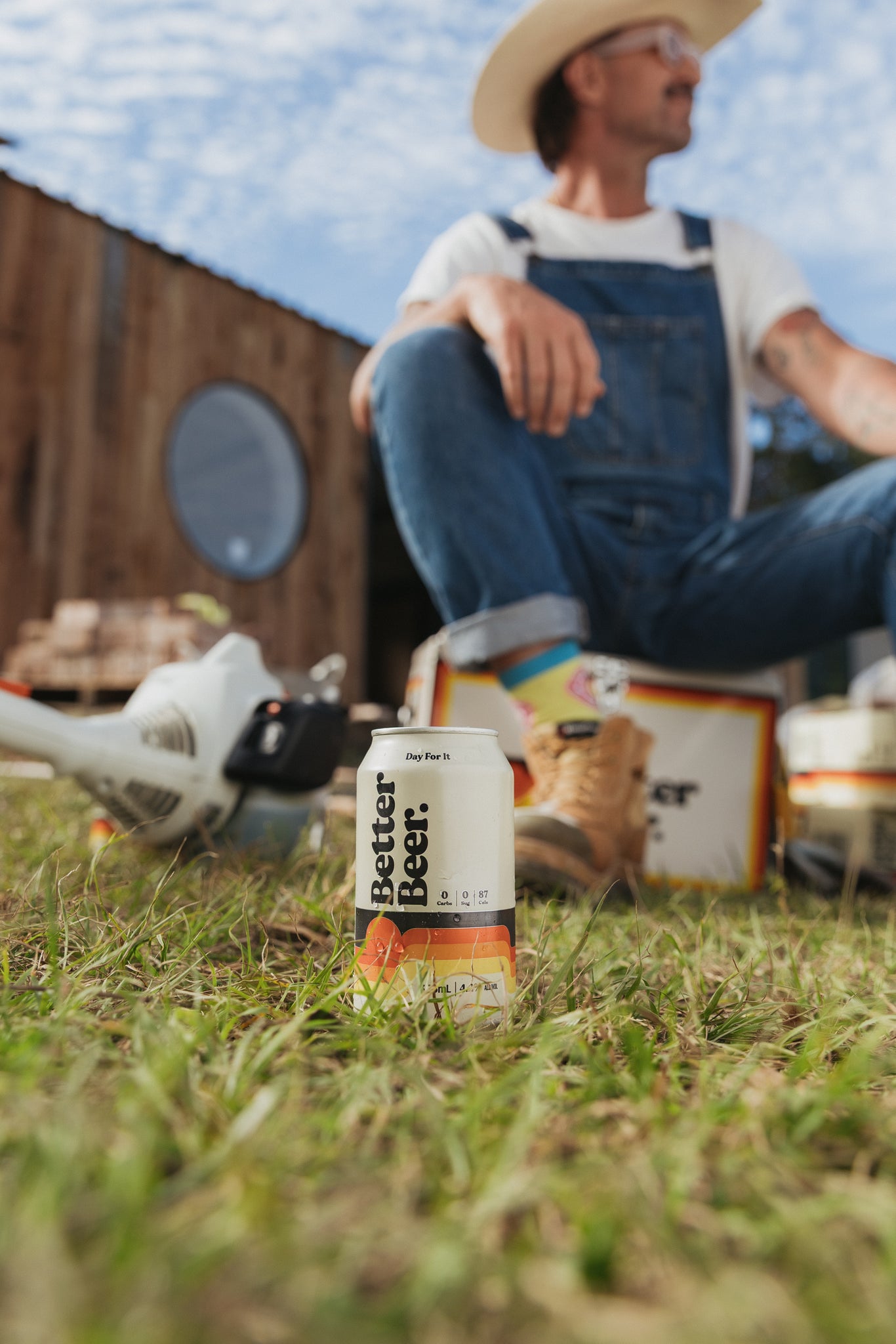 Person in overalls sitting on grass with a can of 'Better Beer' in the foreground.