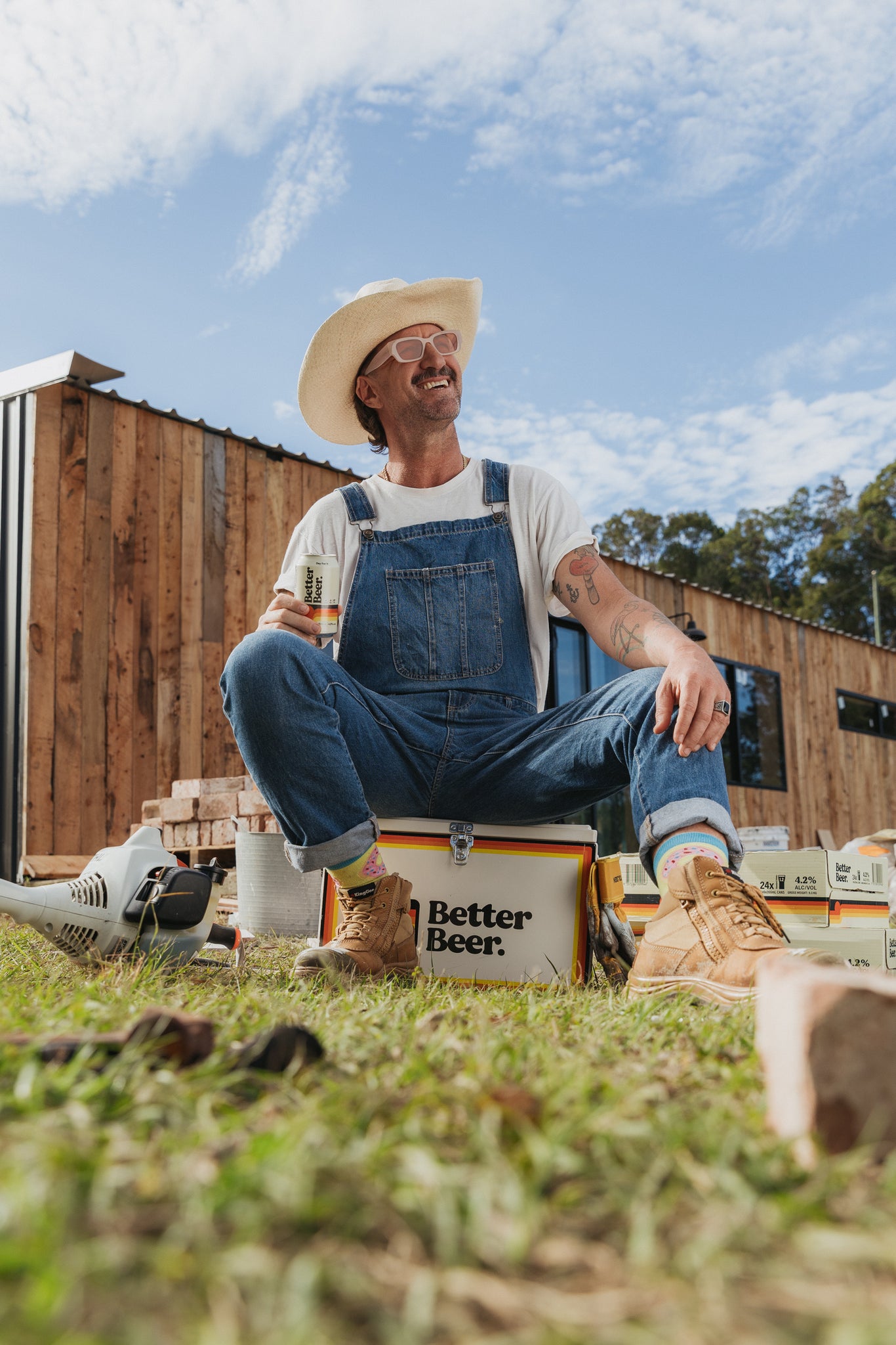Man in overalls and cowboy hat sitting on a 'Better Beer' crate outdoors.