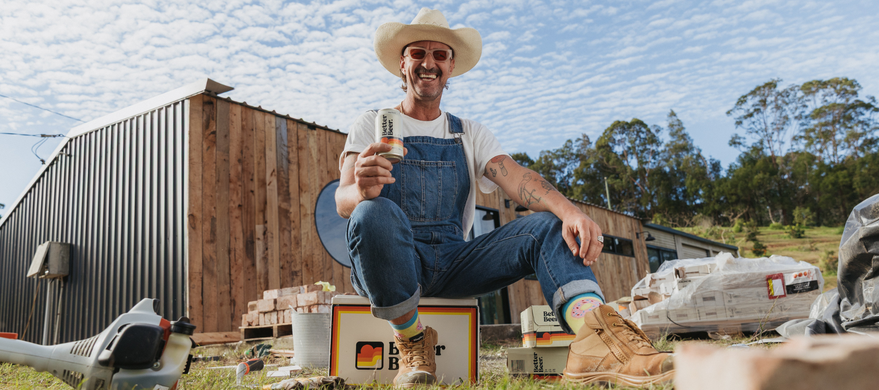 Man in overalls sitting on a esky with a barn and trees in the background