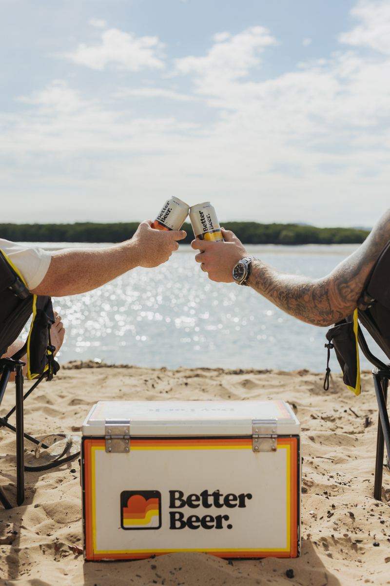 Two people toasting with cans of beer in front of a 'Better Beer' cooler on a beach.
